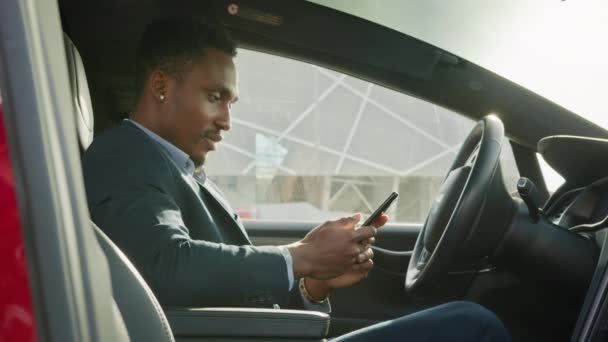 Zijaanzicht van Afrikaanse zakenman in stijlvolle pak rijden auto en spreekt per video in mobiel. Een knappe, drukke man die dringende problemen oplost op weg naar zijn werk. Afrikaanse zakenman aan het praten op mobiel — Stockvideo