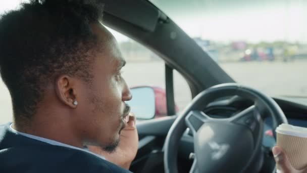 スタイリッシュなスーツ姿のビジネスマンが、コーヒーを片手に高級車の中で座っている。アフリカの男性は仕事上の問題を解決するためにドライブ中に停止します。携帯電話で話してる男 — ストック動画