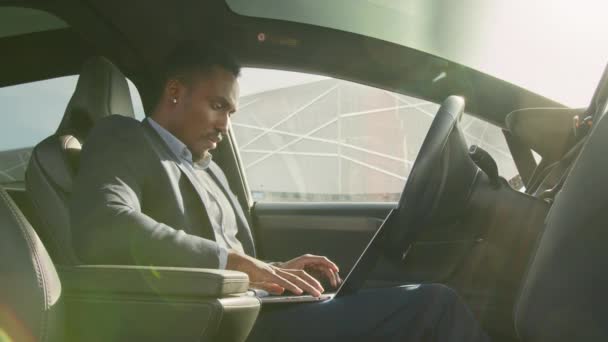 身穿西装的非裔美国人坐在现代电动汽车的司机座位上，用无线笔记本电脑打字。带着手提电脑的忙碌的人。坐在电动汽车上使用笔记本电脑的人 — 图库视频影像