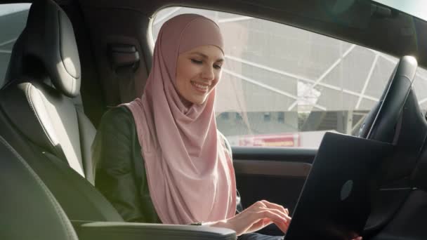 Senhora de negócios atraente no hijab sentado no carro com laptop de joelhos e sorrindo na câmera. Mulher muçulmana trabalhando em computador portátil dentro de veículo de luxo. Senhora no hijab sentado no carro com laptop — Vídeo de Stock