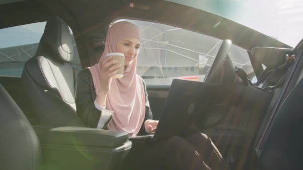 Senhora de negócios atraente no hijab sentado no carro com laptop de joelhos e sorrindo na câmera. Mulher muçulmana trabalhando em computador portátil dentro de veículo de luxo. Senhora sentada dentro do carro elétrico moderno — Vídeo de Stock