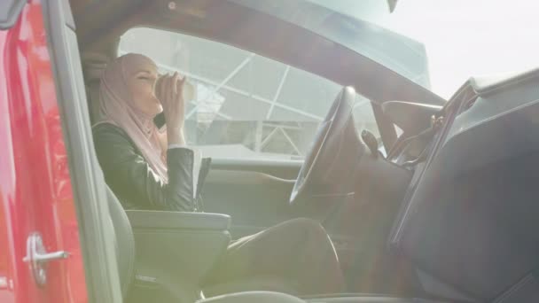 穿着头巾的快乐的年轻女人坐在豪华电动车里一边一边一边用手机一边喝咖啡。在远方工作的女商人人和车辆的概念。戴头巾说话的女人 — 图库视频影像