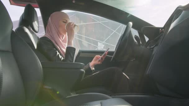 Lüks elektrikli arabasında otururken tesettürlü genç bir kadın telefona bakıyor ve kahve içiyor. Uzakta çalışan bir iş kadını. İnsan ve araç kavramı. Telefon onun elinde. — Stok video