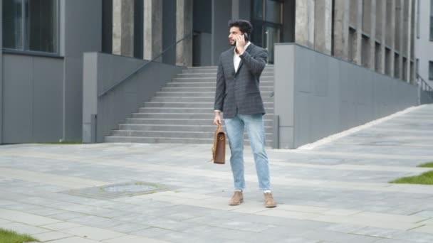 Úspěšný hinduistický podnikatel ve stylovém oblečení stojí v blízkosti kancelářského centra a mluví na mobilu. Hezký muž drží kufr a pomocí moderního smartphonu pro konverzaci na ulici. — Stock video