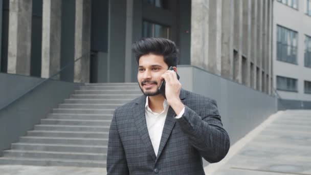 Portrét pozitivního arabského muže ve formálním oblečení, jak vede mobilní konverzaci, když stojí na ulici. Rozmazané pozadí moderní kancelářské budovy. Pojem podnikání a technologie. Portrét muže — Stock video