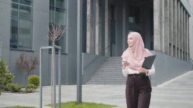 Modern ofis binasının yanında elinde akıllı telefonuyla duran güzel Müslüman kadının alçak açılı görüntüsü. Resmi giyimli ve tesettürlü genç iş kadını dışarıda iş e-postalarını kontrol ediyor. Müslüman iş kadını