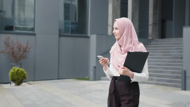 身穿正式服装、头戴头巾的穆斯林女商人站在街上，用手机交谈。有自信的女企业家，手握剪贴板，在智能手机上进行工作对话. — 图库视频影像