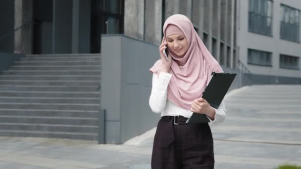 Empreendedora mulher confiante com área de transferência nas mãos tendo uma conversa de trabalho no smartphone. Mulher de negócios muçulmana em roupas formais e hijab em pé na rua e falando no celular. — Vídeo de Stock