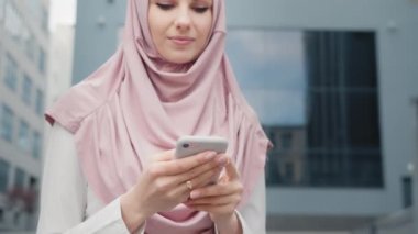 Modern ofis binasının yanında elinde akıllı telefonuyla duran güzel Müslüman kadının alçak açılı görüntüsü. Resmi giyimli ve tesettürlü genç iş kadını dışarıda iş e-postalarını kontrol ediyor. Müslüman iş kadını