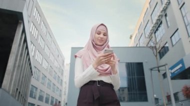 Modern ofis binasının yanında elinde akıllı telefonuyla duran güzel Müslüman kadının alçak açılı görüntüsü. Ofis merkezinde cep telefonu olan Müslüman bir iş kadını. Telefonu olan kız gülümsüyor.