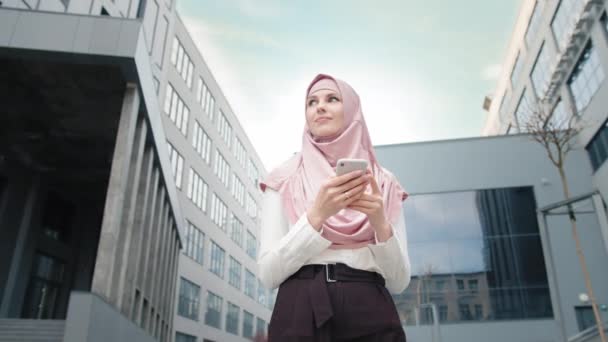 Muslimische Geschäftsfrau mit Mobiltelefon in der Nähe des Bürozentrums. Niedrigwinkel-Ansicht der schönen muslimischen Frau, die in der Nähe moderner Bürogebäude mit Smartphone in der Hand steht.Das Mädchen mit dem Telefon lächelt — Stockvideo