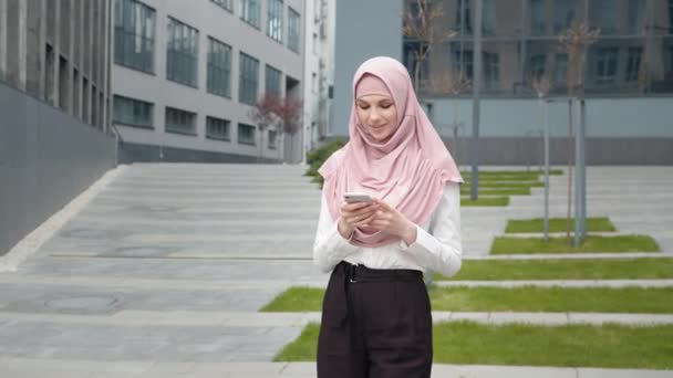 Närbild av trevlig affärskvinna i hijab med modern smartphone när du står utomhus. Unga arabier skriver meddelanden på mobilen utanför kontoret. Affärskvinna stående nära kontorshotell — Stockvideo