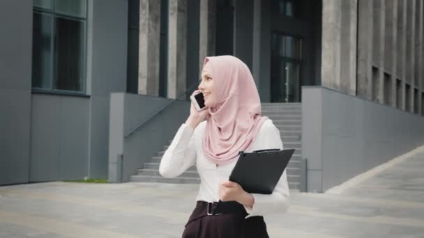 Retrato de mulher árabe no hijab em pé perto do moderno centro de negócios com smartphone e área de transferência nas mãos. Mulher de negócios muçulmana em roupas formais e hijab passa na rua e fala no celular — Vídeo de Stock