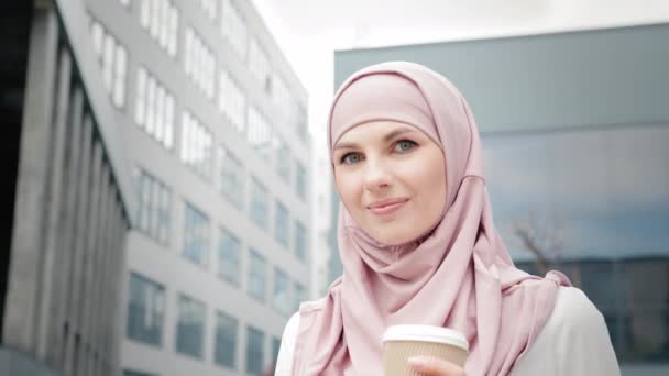 Türbanlı genç bir müslüman kızın portresi. Arap iş kadını tesettürlü, kahve içen ve dışarıda ofis binasının yanında dururken akıllı telefon kullanan. Teknoloji, iş ve modern yaşam tarzları kavramı — Stok video