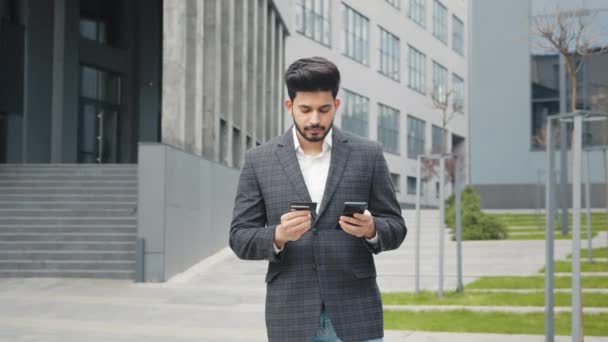 Bonito homem hindu em roupas formais elegantes usando smartphone moderno e cartão de crédito para compras online. Homem de negócios barbudo na rua e a fazer compras. Homem hindu de terno usando smartphone — Vídeo de Stock