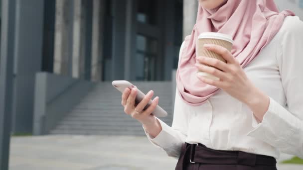 站在商业中心附近的穆斯林妇女手持现代智能手机和外卖咖啡，站在伊斯兰头巾的近旁。背景模糊。成功的女性职业。一个穿着头巾的年轻穆斯林女孩的画像 — 图库视频影像