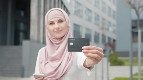 Donna d'affari in hijab con carta di credito e smartphone fare acquisti all'aperto. Sfocatura sfondo di donne. Concentrati sulle mani femminili che tengono la carta bancaria nera. Donna in possesso di smartphone e carta di credito — Video Stock