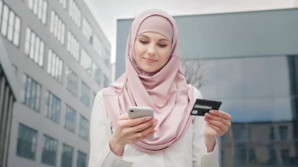 Kredi kartı olan Arap bir kadın. Resmi giyinmiş, tesettürlü güzel Müslüman kadın kredi kartı ve akıllı telefonuyla online alışveriş yapıyor. Bayan ofis binasının yanında duruyor ve kolay ödemenin keyfini çıkarıyor. — Stok video