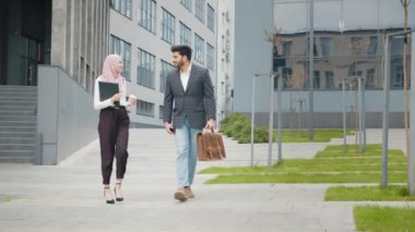 Sırıtan tesettürlü kadın bavullu müslüman adamla ofis binasının yanında yürürken pano ve kahve taşıyor. İki iş ortağı sokakta ortak projeyi tartışıyor. İki ofis çalışanı