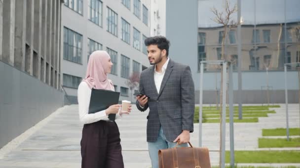 Hombre de negocios musulmán en traje mostrando algo en el teléfono inteligente a su colega en hiyab. Mujer bonita de pie cerca y sosteniendo en las manos portapapeles y taza de café. Hombre mostrando en el móvil — Vídeos de Stock