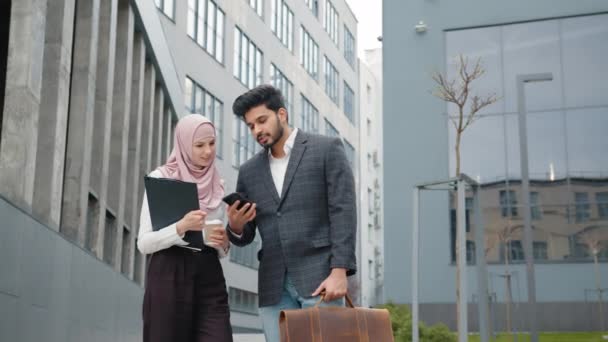 Compañeros de negocios árabes en ropa formal caminando por la calle y discutiendo algunos temas de trabajo. Hombre joven con maleta usando smartphone, mujer en hijab llevando portapapeles. Compañeros de negocios — Vídeos de Stock
