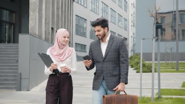 Arap iş arkadaşları resmi giysiler içinde sokakta yürüyor ve bazı iş konularını tartışıyorlar. Takım elbiseli yakışıklı bir adam bavul ve akıllı telefon taşıyor, tesettürlü çekici bir kadın elinde kahve tutuyor. — Stok video