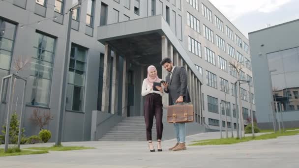 Μουσουλμάνος επιχειρηματίας με κοστούμι που δείχνει κάτι στο smartphone στη γυναίκα συνάδελφό του με μαντίλα. Αραβες συνάδελφοι επιχειρηματίες με επίσημα ρούχα περπατούν στο δρόμο και συζητούν κάποιο θέμα εργασίας — Αρχείο Βίντεο