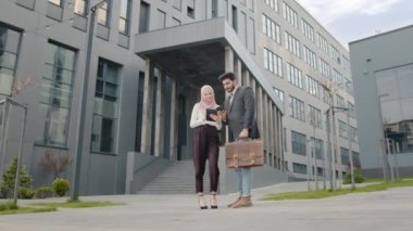 İş arkadaşları iş hakkında konuşuyor. Dijital tabletli tesettürlü bir kadın ve akıllı telefonlu Müslüman bir adam ofis binasının yakınında yan yana duruyorlar. İki meslektaş dışarıda çalışmak için modern aletler kullanıyor..
