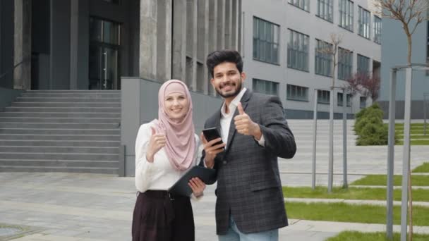 Mujer sonriente en hijab y musulmán hombre guapo mostrando los pulgares hacia arriba mientras que de pie al aire libre cerca del edificio de oficinas. Cooperación exitosa de dos socios comerciales. Arabian business people showing thumbs. — Vídeos de Stock