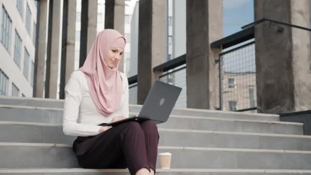 Porträtt av attraktiv arabisk kvinna i eleganta kläder och hijab sitter på trappor med modern bärbar dator på knä. Ung kvinna som arbetar på bärbar dator utomhus. Kvinna som sitter på trappor med bärbar dator — Stockvideo