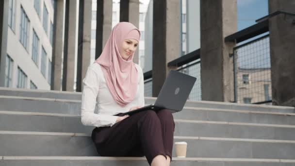 Retrato de mujer árabe atractiva en ropa elegante y hijab sentado en las escaleras con un portátil moderno en las rodillas. Encantadora mujer en ropa elegante con estilo y hijab. Mujer musulmana trabajando a distancia — Vídeo de stock