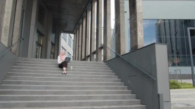 Genç Müslüman kadın merdivenlerde iş merkezinin yanında oturuyor ve kablosuz bilgisayarla çalışıyor. Zarif kıyafetler ve tesettürlü güzel bir kadın. Serbest meslek kavramı. Merdivenlerde oturan ve dizüstü bilgisayar kullanan kadın.