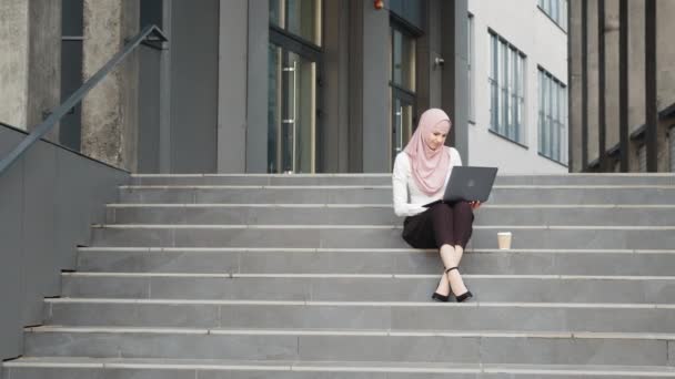 Młoda muzułmanka siedząca na schodach w pobliżu centrum biznesowego i pracująca na bezprzewodowym laptopie. Ładna kobieta ubrana w eleganckie ubrania i hidżab. Koncepcja wolnego strzelca. Muzułmanka pracująca na laptopie na zewnątrz — Wideo stockowe