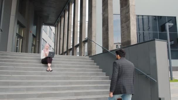 Vista lateral do homem árabe em roupas elegantes vai em escadas com mala nas mãos. Mulher encantadora em hijab trabalhando em laptop no fundo. Dois colegas de trabalho ao ar livre. Mulher muçulmana trabalhando no laptop — Vídeo de Stock