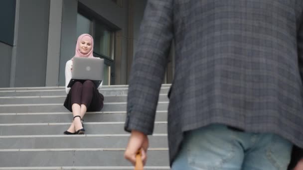 Mulher sorridente em hijab e desgaste formal sentado com laptop moderno em joelhos. Feche-se o homem com a mala a subir as escadas. Dois sócios a trabalhar ao ar livre. Parceiros de negócios que têm reunião ao ar livre — Vídeo de Stock