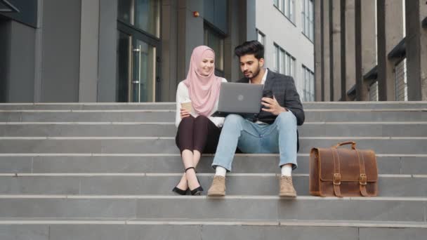 Pria muslim bergaya dan wanita menawan mengenakan jilbab duduk bersama di tangga, tersenyum dan berbicara. Dua pekerja lepas menggunakan laptop untuk bekerja di luar ruangan. Dua rekan arab duduk di tangga — Stok Video