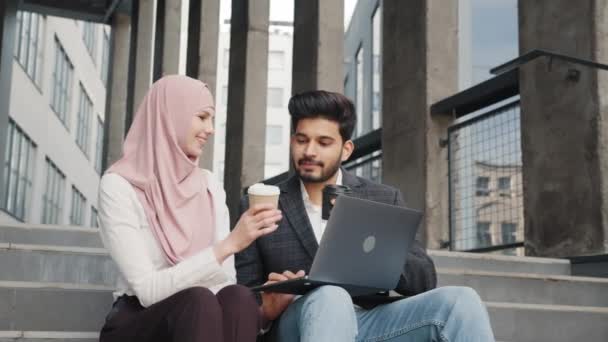 Socios árabes que utilizan el ordenador portátil para trabajar al aire libre portátil. Feliz musulmán hombre y mujer tostadas con tazas de café mientras se sientan juntos en las escaleras al aire libre. Socios comerciales que utilizan ordenador portátil para reuniones — Vídeos de Stock