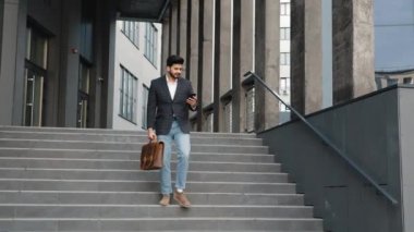 Bir elinde modern akıllı telefon, diğerinde deri kahverengi bavul ile merdivenlerden inen Arap adamın tam boy portresi. İş anlayışı, başarı ve güven. Arap adam.