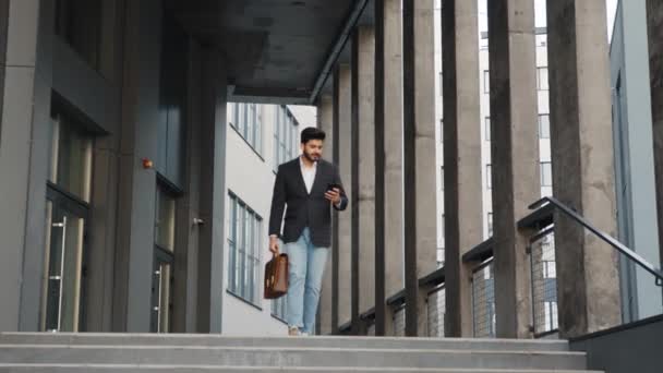 Retrato de comprimento total do homem árabe descendo escadas com smartphone moderno em uma mão e mala marrom de couro em outra. Conceito de negócio, sucesso e confiança. gadget moderno para o trabalho — Vídeo de Stock