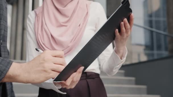 Wanita mengenakan jilbab memegang papan klip sementara pria menandatangani kontrak. Pria Arab menandatangani surat-surat di clipboard bahwa wanita memegang. Rekan bisnis dalam pakaian formal berdiri di tangga di luar kantor — Stok Video