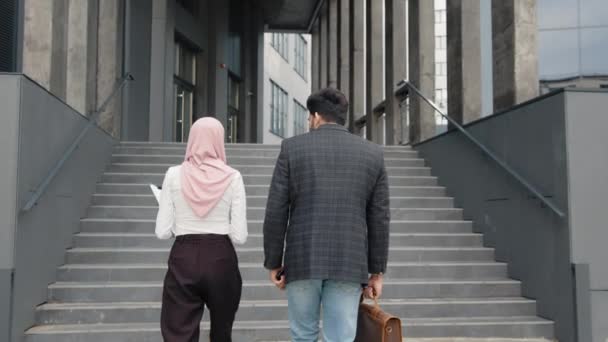 Modern ofis binasının yakınında yürüyen Müslüman kadın ve erkek portresi çalışma anlarını tartışıyor. Açık havada akıllı telefonları, panosu ve bavulu olan iki iş arkadaşı. Sırtından vurdum. — Stok video