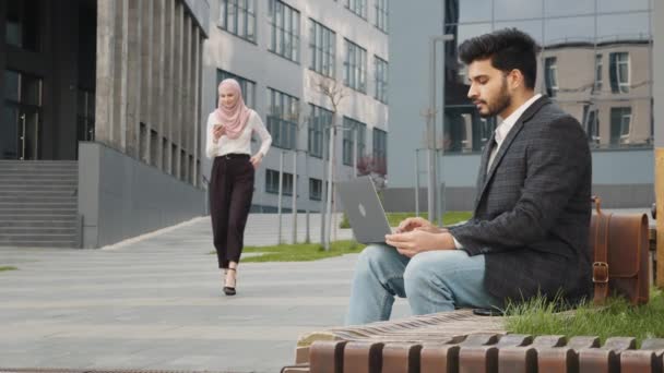Rozmazané pozadí muslimského muže ve stylovém oblečení. Hezká žena v hidžábu kráčí na pozadí s chytrým telefonem na rukou. Městská oblast. Arabský muž pracuje na notebooku, zatímco žena v hidžábu na mobilu — Stock video