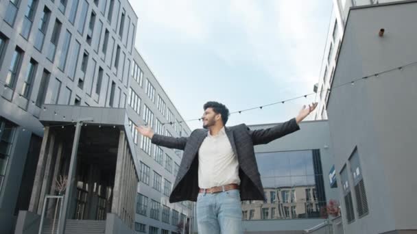 Vista de ángulo bajo del hombre de negocios hindú guapo de pie cerca del moderno edificio de oficinas con las manos extendidas y sintiendo felicidad. Concepto de emociones humanas y carrera de éxito. Feliz hombre de negocios hindú — Vídeos de Stock