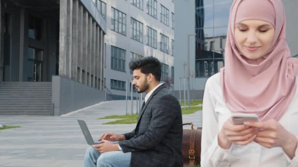 Trevlig kvinna i hijab sitter på träbänk utomhus och håller modern mobiltelefon. Oskärpa bakgrund av arabier man som arbetar på trådlös bärbar dator. Begreppet affärsmän och prylar. — Stockvideo