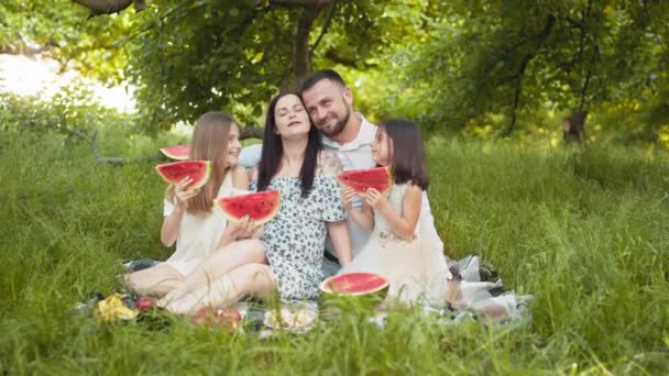 Padres jóvenes positivos con dos hijas pequeñas comiendo sandía durante el picnic al aire libre. Familia feliz relajándose juntos en la naturaleza durante la temporada de verano. Padres con niños comiendo sandía — Vídeo de stock