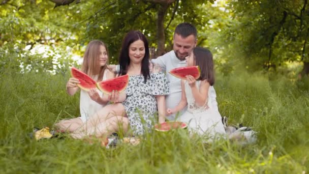 Vrolijke ouders met dochters in zomerse outfits zitten samen in de groene tuin en genieten van zoete watermeloen. Een picknick buiten. Ontspanningstijd. Ouders met kinderen picknicken met zoet fruit — Stockvideo