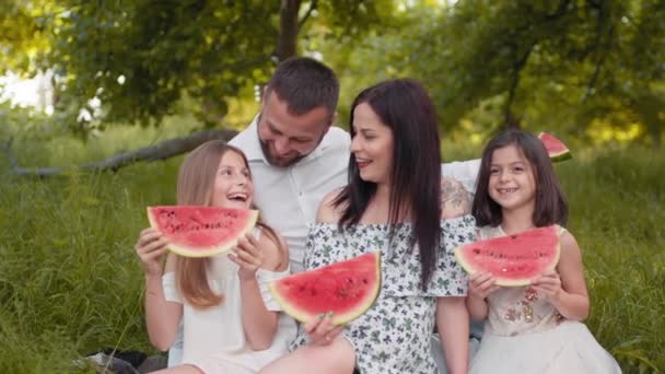 Happy family with kids having summer picnic at green park. Young parents and two daughters eating sweet watermelon. Outdoors relaxation and enjoyment. Parents with two kids eating watermelon at picnic — Stock Video