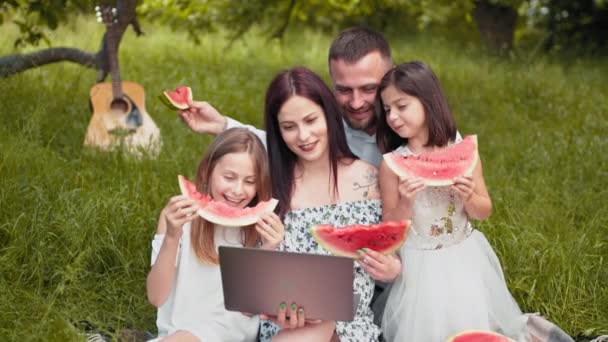 Familia caucásica positiva haciendo picnic de verano con frutas dulces y cupcakes. Mujer sosteniendo portátil inalámbrico mientras dos hijas y marido la abrazan. Tener videollamada en el portátil durante el picnic — Vídeo de stock
