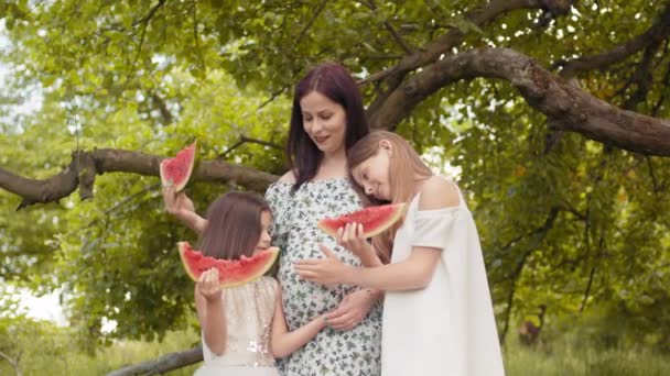 Mujer caucásica embarazada y sus dos hermosas hijas de pie juntas en abrazos en el jardín verde. Familia feliz comiendo sandía dulce al aire libre. Dos niñas tocando las madres embarazadas vientre en el jardín — Vídeo de stock