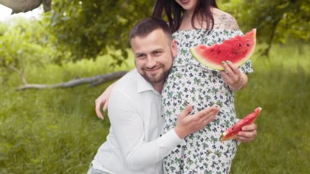 Gelukkig getrouwd stel met watermeloen in handen poserend bij de groene tuin. Knappe man die een zwangere vrouw omarmt. Familie en liefde concept. Knappe man omhelst zwangere vrouw buiten — Stockvideo